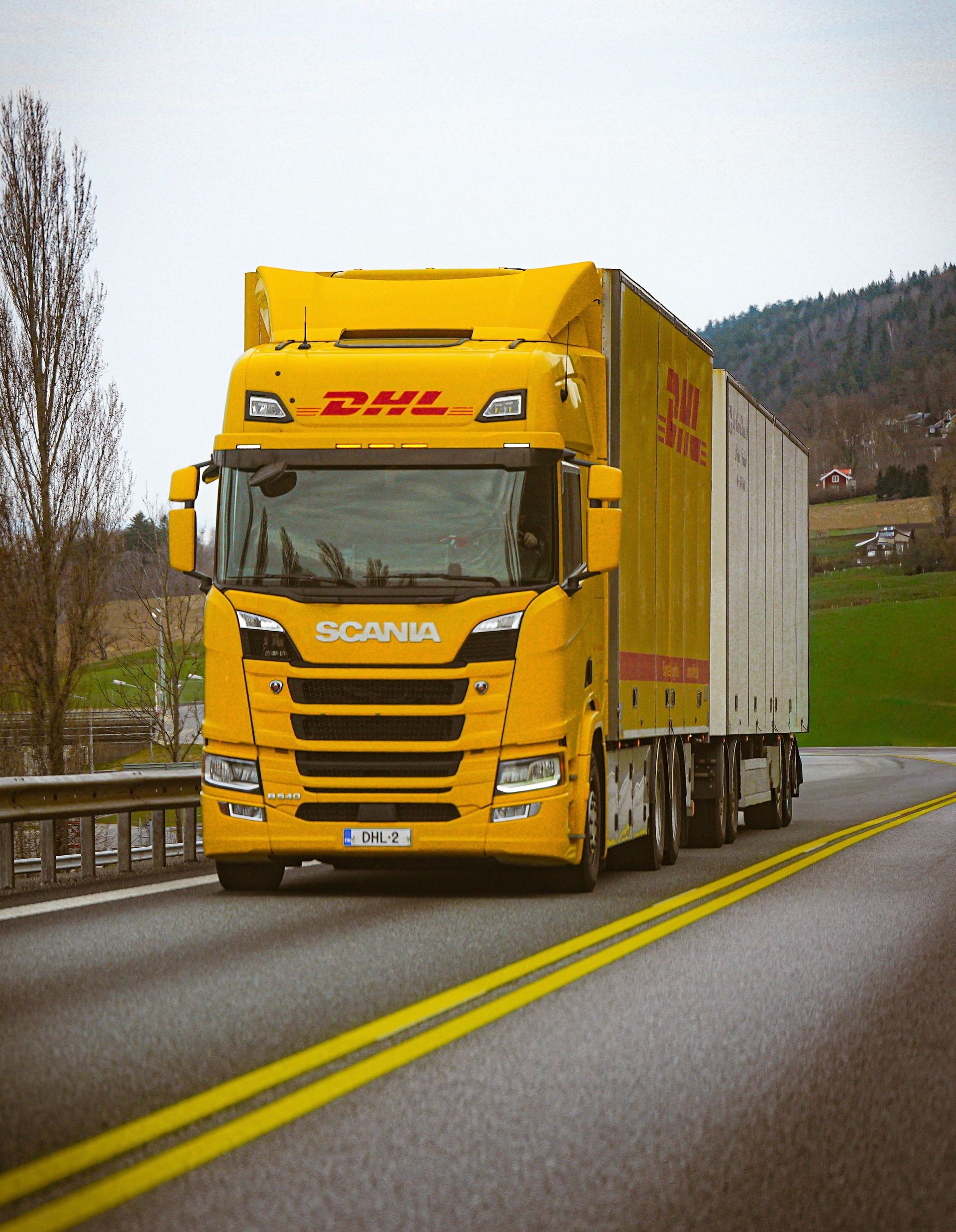 A yellow DHL truck on a scenic highway with green hills in the background.