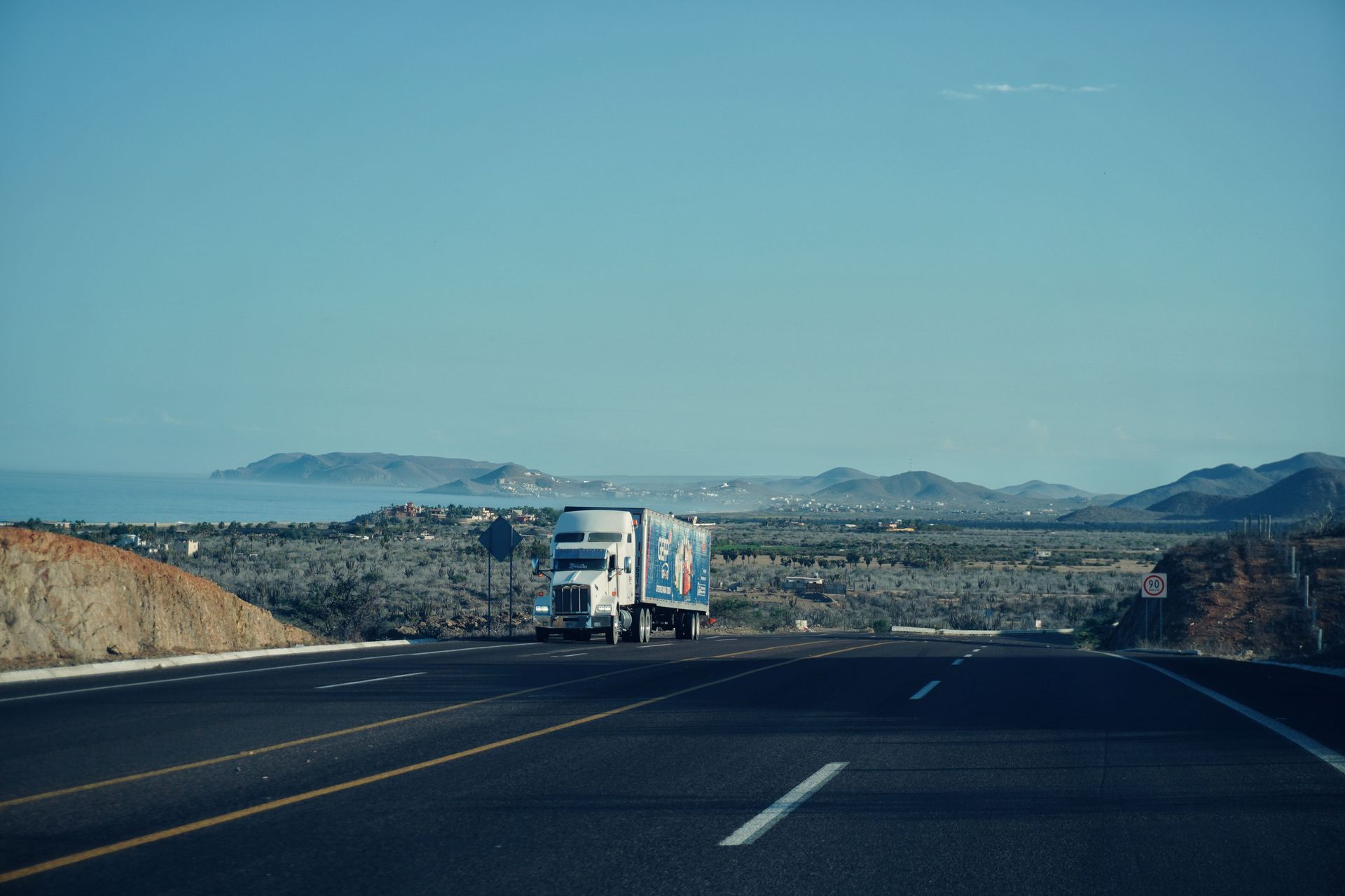 A semi-truck travels on a scenic highway with mountains in the background under a clear sky.